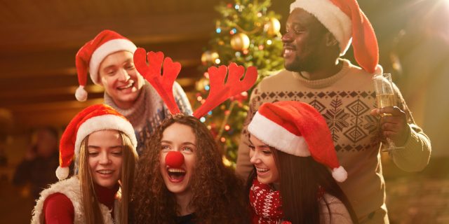 A group of friends taking a selfie in front of the Christmas tree, holding gifts and wearing antler headbands and Santa hats.