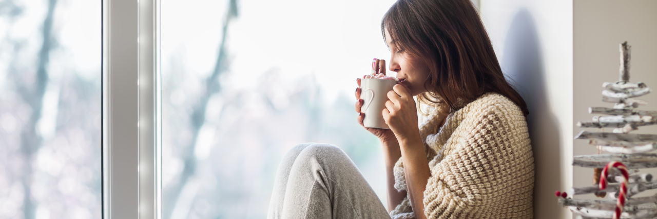 Mental Health Self-Care Tips for the Upcoming Holiday Season Young beautiful woman drinking hot coffee sitting on window sill