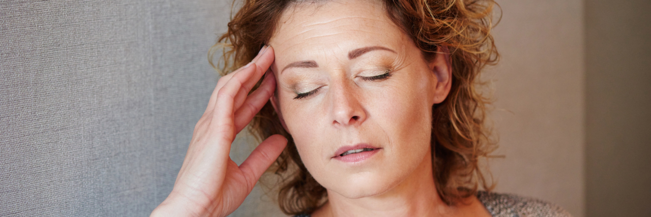 What to Remember When You're Experiencing Fibromyalgia Fatigue A middle-aged woman with her hand in her head, with her eyes closed. Looking overwhelmed and tired.