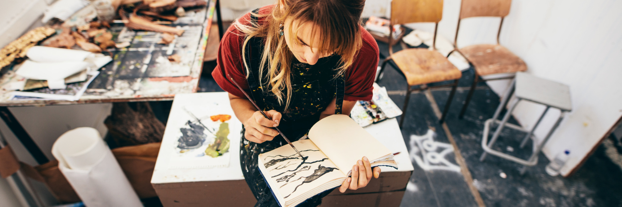 9 Gift Ideas for the Creative Spirit Who Struggles With Mental Illness Female artist drawing pictures in her workshop. Young woman sitting and painting in her drawing book.
