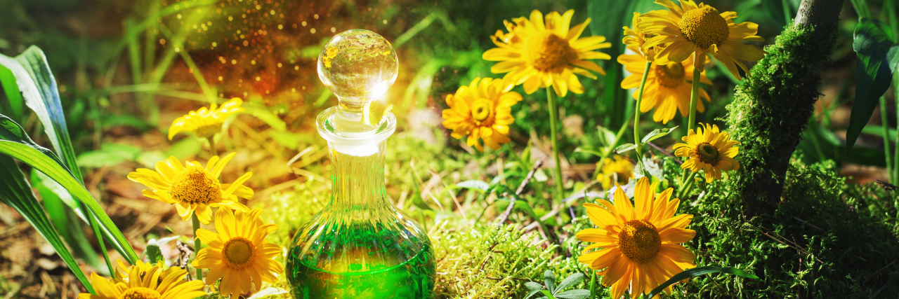 A bottle filled with green liquid, sitting in some grass, surrounded by yellow flowers with the sun shining down on it.