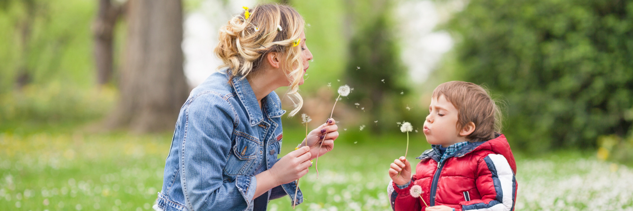 A mom and her young son blowing on dandelions in a park.