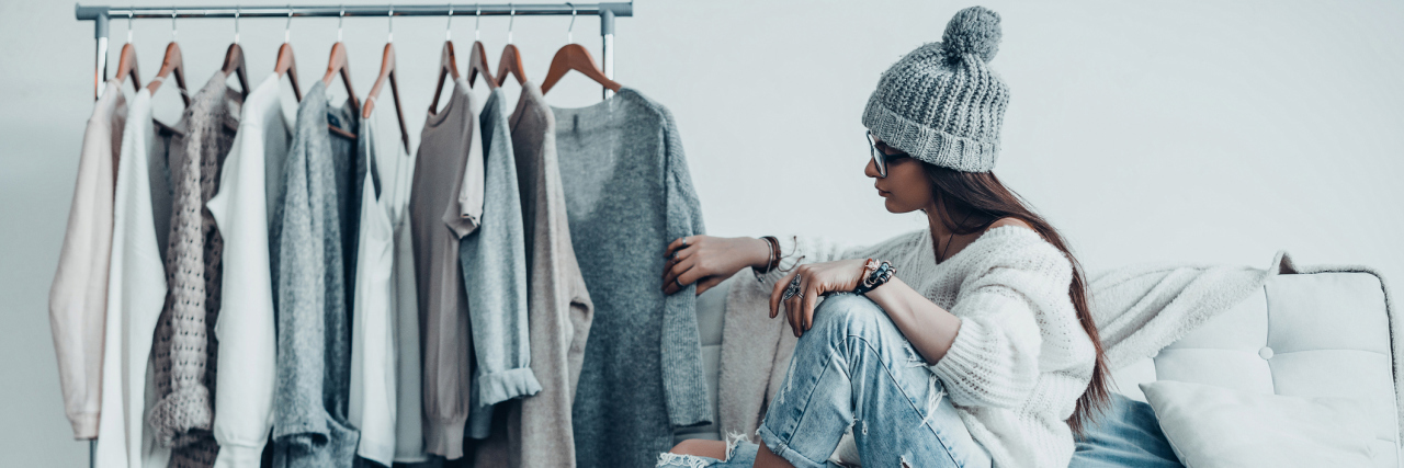 4 Ways My Illnesses Affect What I Wear A woman sitting on a chair, looking at a rack of clothing.