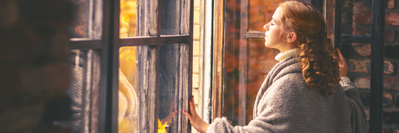 What It's Like to Be Chronically Ill in the Fall A woman looking outside her window, out into the fall.
