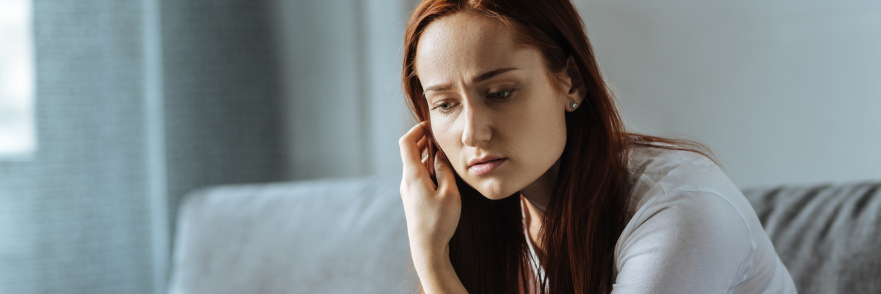 Receiving the Needed Validation for My Health Came at a Cost A young woman sitting on a couch, with a serious expression, in deep thought.