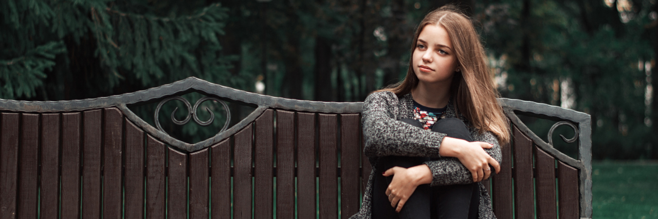 The Reality of Sensory Processing Disorder in Teens Teenage girl with blond hair in knitted cardigan and black denim jeans sitting on wooden bench in the park.