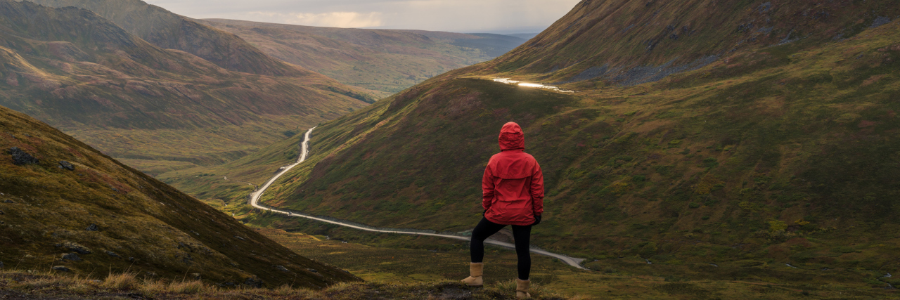 The Difficulties of Learning to Live With Fibromyalgia woman in a red jacket standing on top of a mountain looking down at small winding path
