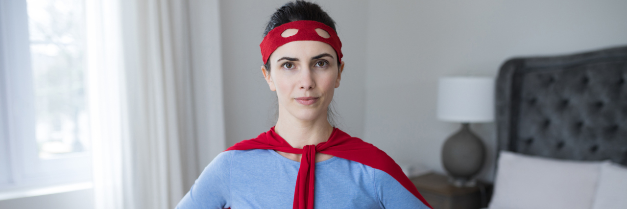 Why I Think People With Chronic Illnesses Are Superheroes A woman standing in her bedroom, wearing a cape and superhero mask over her eyes.