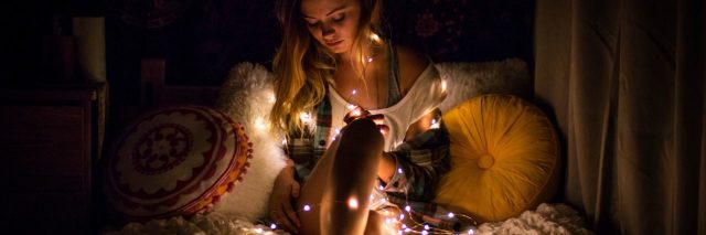 young blonde woman sitting on bed surrounded by fairy lights