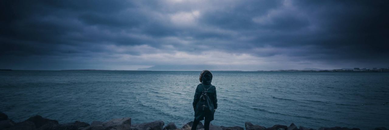 The Importance of Self-Care in the Wake of Natural Disasters woman in coat standing in front of stormy skies and the ocean
