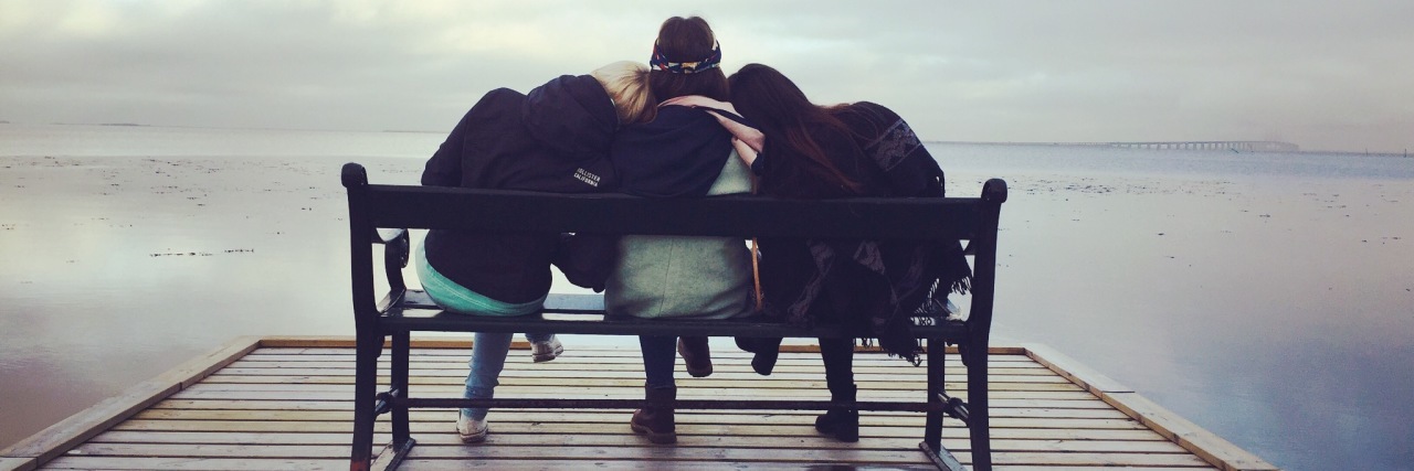 three friends sitting on bench facing end of dock over water