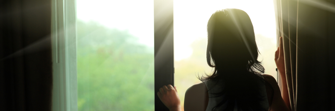 How I Stay Optimistic With Chronic Illness woman opening the curtain to look at the sunrise