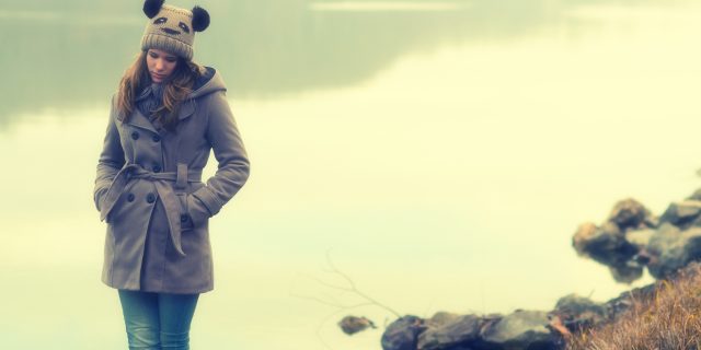 sad young woman standing outside alone next to lake in winter