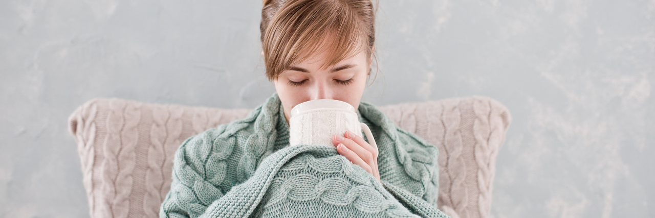 Attractive woman with natural blond hair posing without makeup with white cup of hot drink wrapped with blanket at home