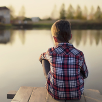 child dreaming sitting on a wooden pier near the water and looking at the house and the people on the other side