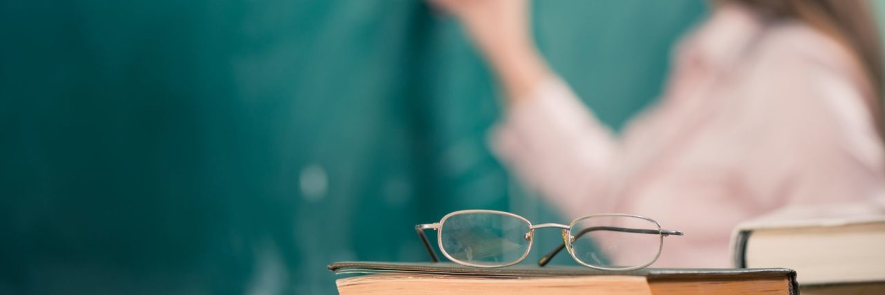 glasses and books on teacher's table in focus with female teacher at background at chalk board