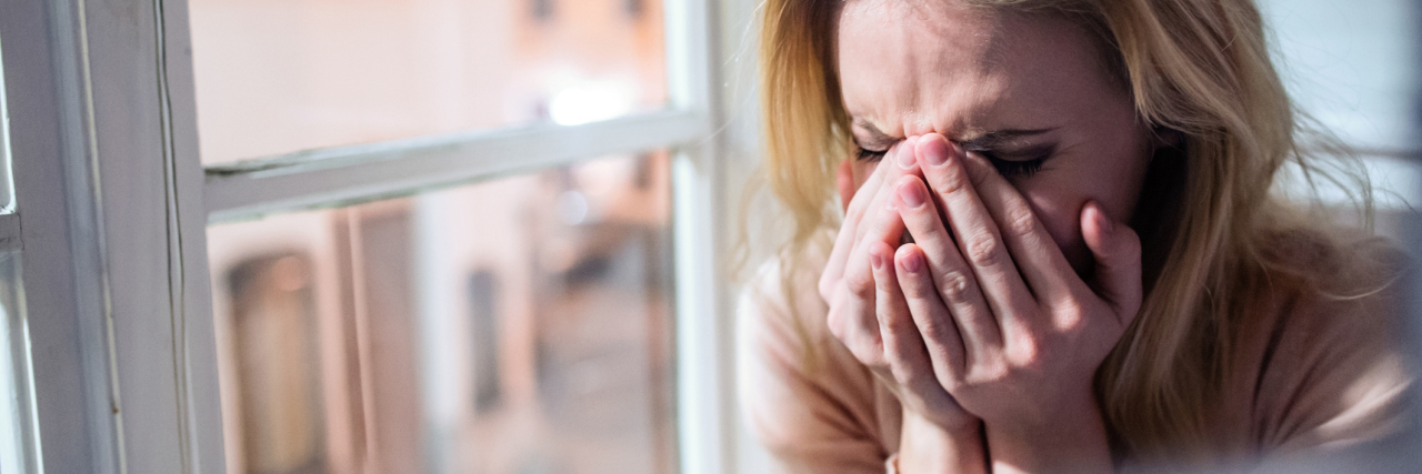 blonde woman crying beside window hands partly covering her face