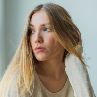 Young blonde woman sitting next to window in sweater, looking emotive and thoughtful