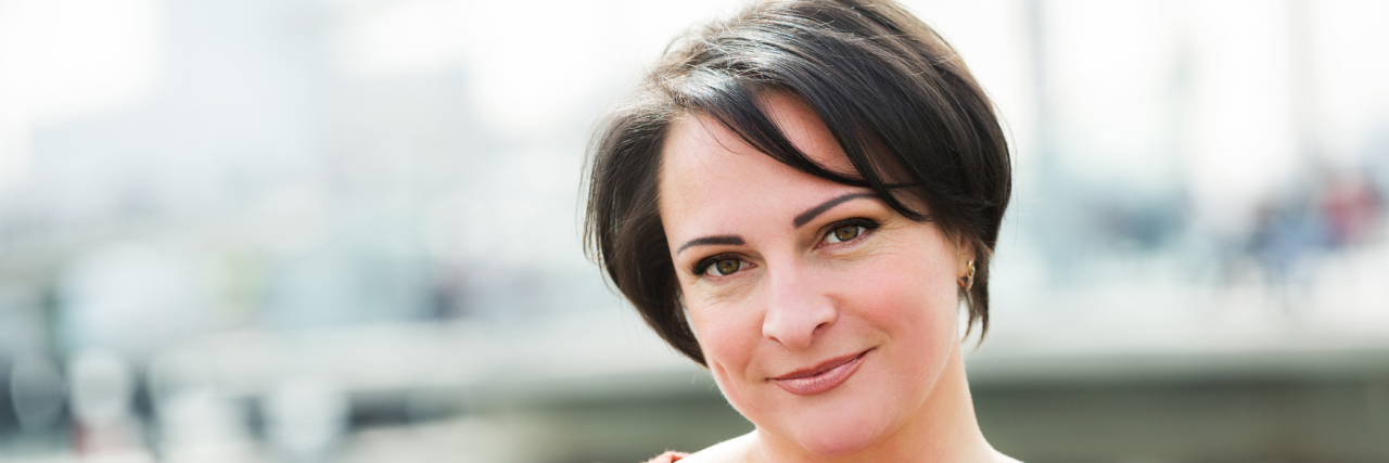 Finding Reasons to Smile in Life With Chronic Pain woman with short brown hair wearing a red shirt standing outside and smiling