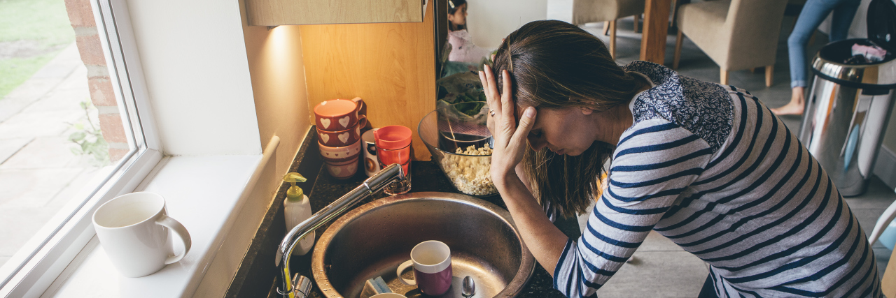 Invisible Struggles of a Mother With Asperger's Syndrome Stressed mum at home. She has her head in her hands at a messy kitchen sink and her children are running round in the background.