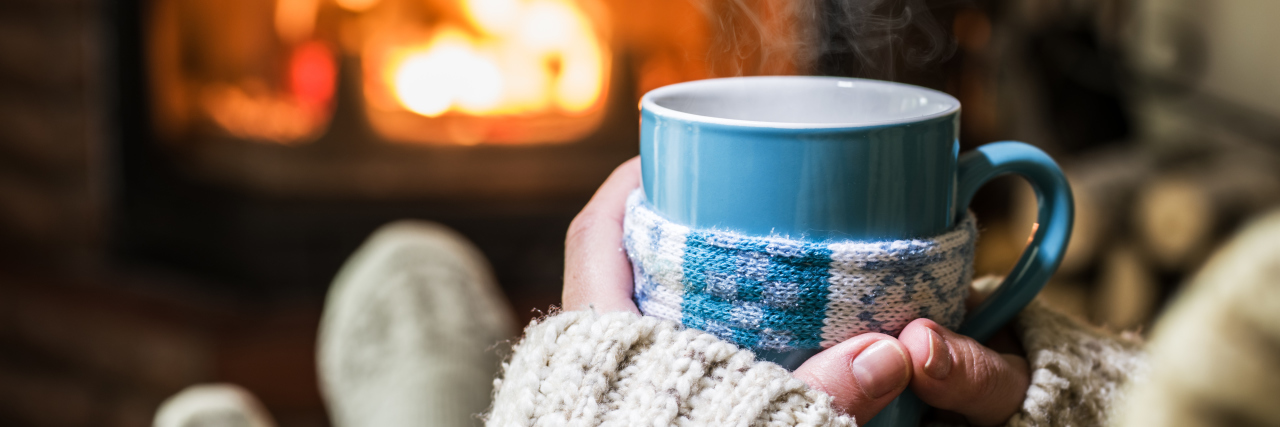 Being Home During Christmas Because of Chronic Illness woman sitting with a mug and a blanket in front of the fire
