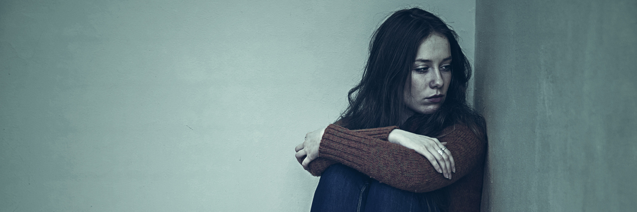 The Guilt That Comes With Endometriosis A woman sitting on the floor, looking at the wall with a solemn facial expression.
