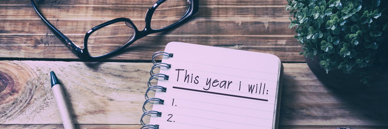Setting New Year's Resolutions While Chronically Ill A notepad on a wooden table, surrounded by a pen, glasses, and greenery.