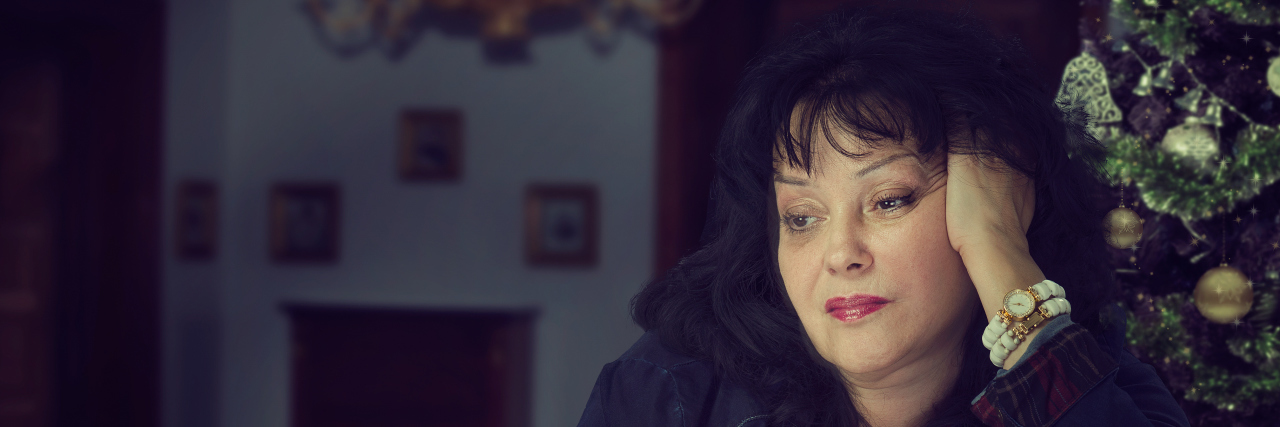 The Truth About Not Having a Biological Family Around the Holidays Black-haired mature woman is upset and frustrated about celebrating Christmas alone. Woman drinks coffee, head is propped up with her hand. Dark blurred indoors background with decorated tree