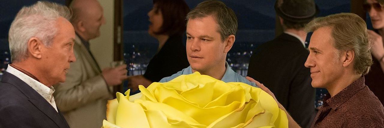 A photo of the movie "Downsizing," with Matt Damon holding a large rose that is bigger than him.