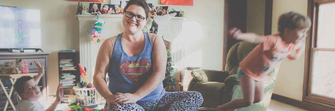 When I Couldn't Pretend Any Longer I Was the 'Perfect' Mom Mother sitting in middle of messy living room, smiling at camera while a child jumps off a couch and one makes a mess on floor