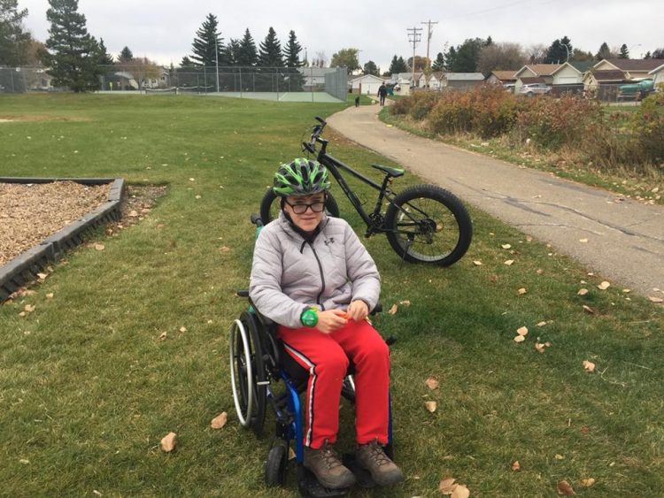 boy wearing a helmet and sitting in a wheelchair at the park with his bike propped up behind him