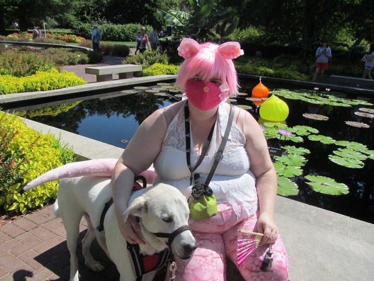 woman with pink hair and a pink face mask sitting in front of a lily pond with her service dog standing next to her