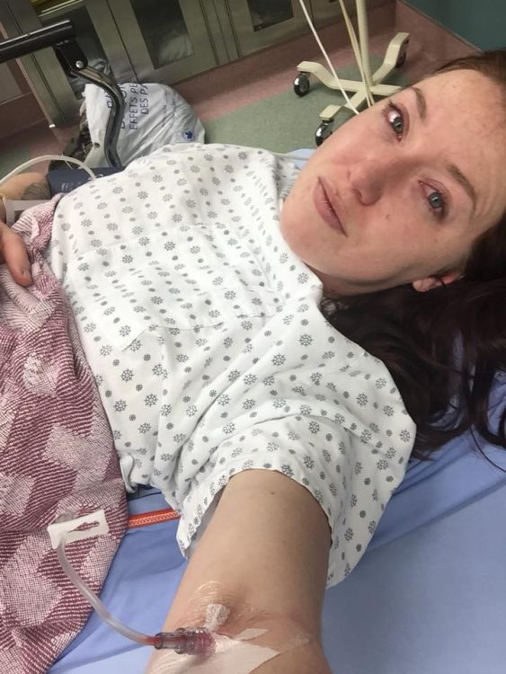 woman taking a selfie in the hospital. she has an IV connected to her arm, is wearing a hospital gown and lying under a blanket