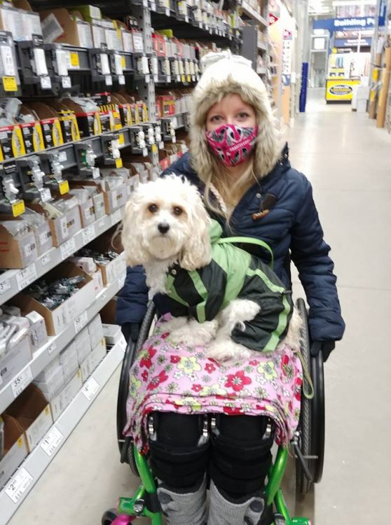 woman sitting in a wheelchair and wearing a facemask in an aisle at lowe's. she has a blanket and a dog in her lap.