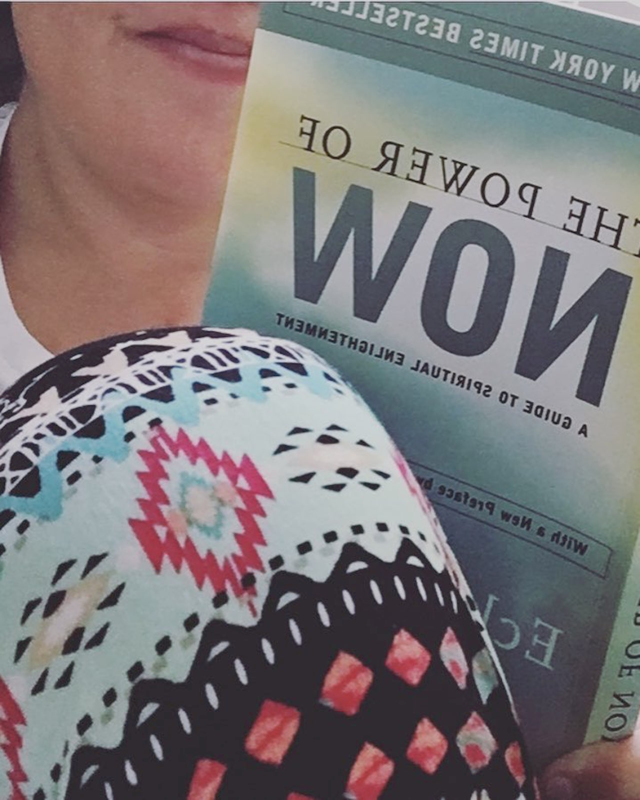 woman wearing patterned leggings and holding a book that says "the power of now"