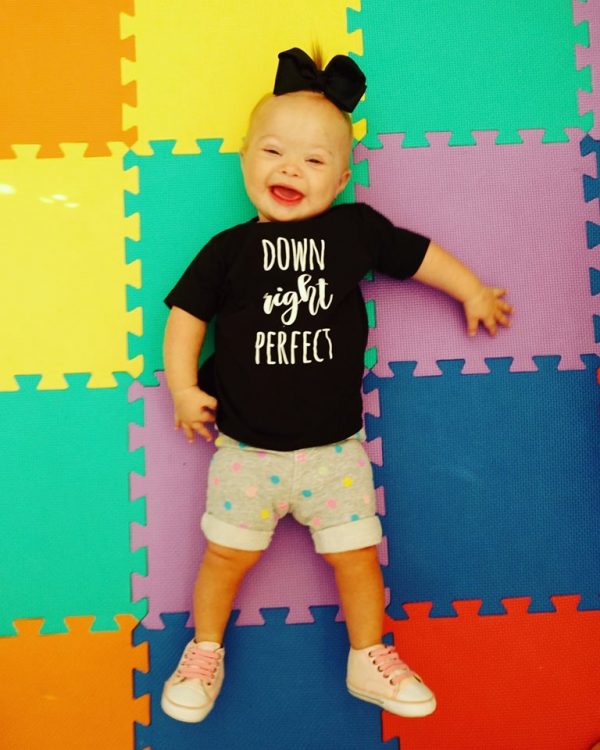 Baby with Down syndrome laying on colorful mat wearing a black shirt tat says "Down right perfect" and black bow.