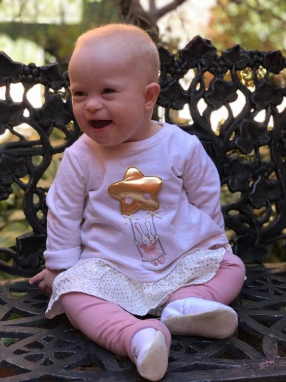 Baby girl with Down syndrome sitting and looking at someone off camera and smiling. She wears all pink and has almost no hair.