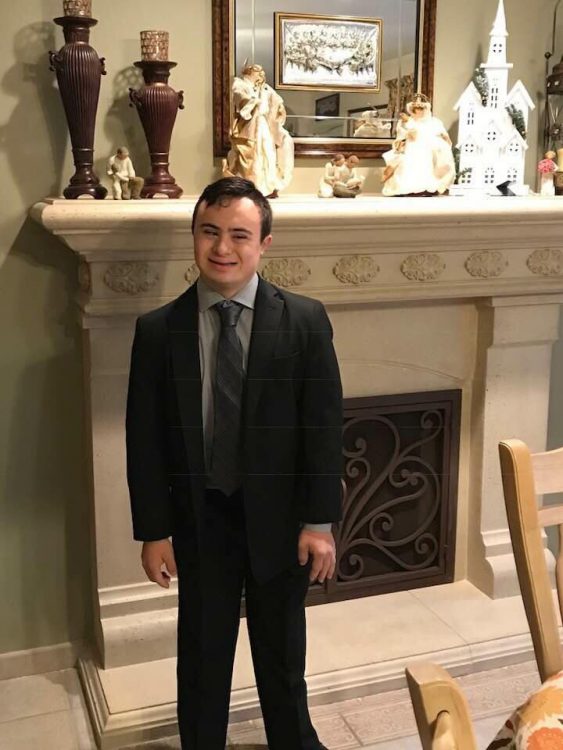 Young man with Down syndrome wearing a suit and posing by a white marble fireplace. 