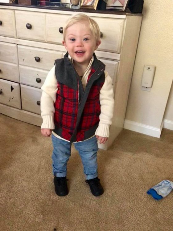 Little boy with Down syndrome posing for the camera, full body shot and he is wearing a white sweater with a red checkered vest and jeans.