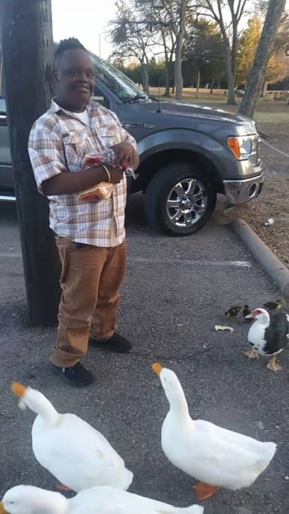 Young teen with Down syndrome standing in a parking lot surrounded by ducks as he feeds them. He is looking at the camera and smiling. 