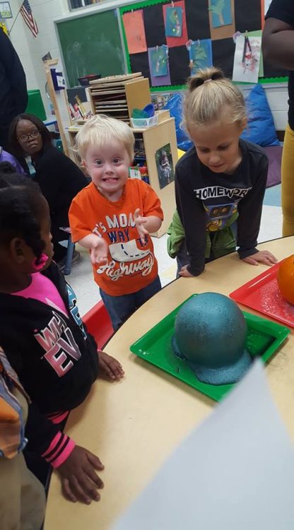 Little boy with Down syndrome at school. He is wearing an orange shirt and looking at the camera with an excited look on his face, big grin and clenching fists.