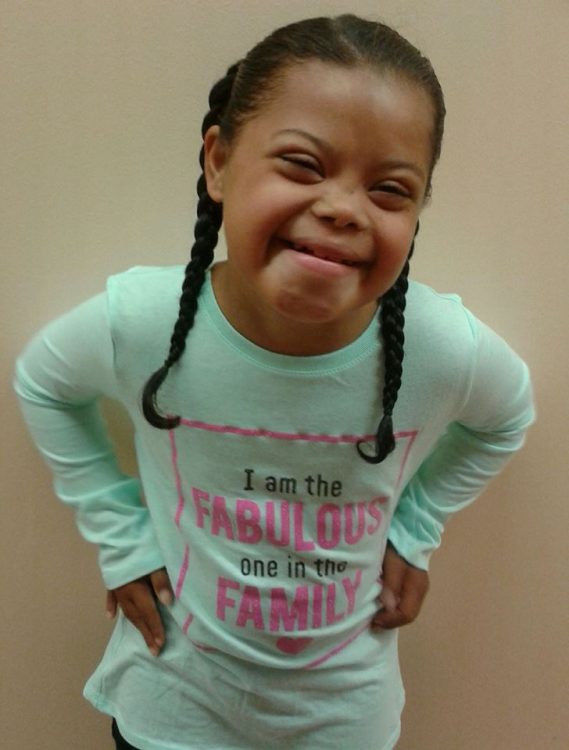 Little girl with Down syndrome posing at camera hands on her hips. She has two braids and a mint colored long sleeved shirt.