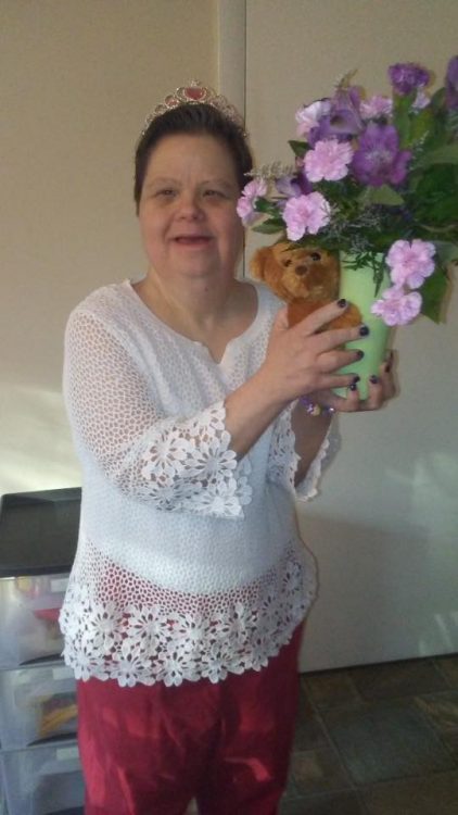 Woman with Down syndrome wearing a tiara, holding a vase of flowers and a teddy bear.