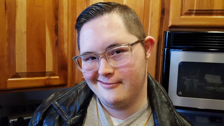 Young adult with Down syndrome smiling at camera. Close up of face, he wears glasses and a leather jacket.