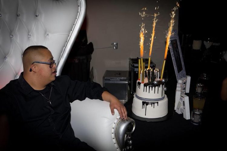 A teenager with Down syndrome sitting on a fancy high back white chair and looking to his cake sitting on a table beside him.