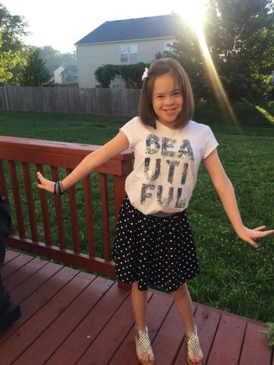 Pre teen with Down syndrome posing for a photo. She wears a pale pink shirt that says, "Beautiful" and a black skirt. She is standing on a red-stained porch.