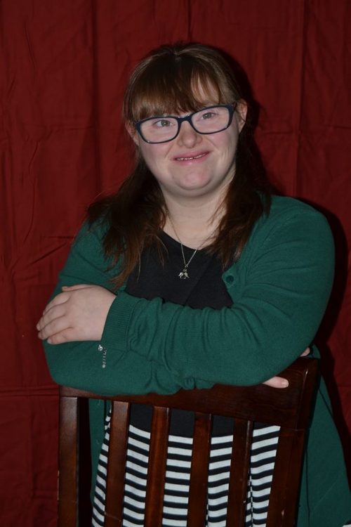 Young woman with Down syndrome looking at the camera with her arms crossed resting on the back of a chair. She has bangs, wears glasses and a green sweater.