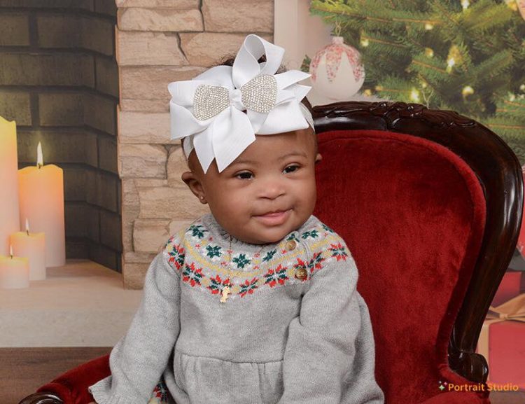 Little girl with Down syndrome wearing a big white bow and sitting on a fancy red velvet chair.