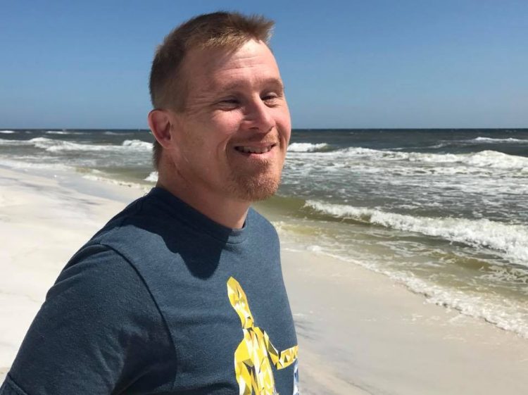 Man with Down syndrome looking at the camera and smiling. He has a goatee and is posing at the beach.