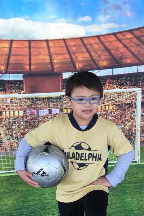 Little boy with Down syndrome wearing a soccer outfit. He is holding the ball and posing in front of the soccer net.